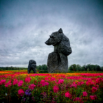 Giant crude carving of a wolf sitting in a field of red flowers.